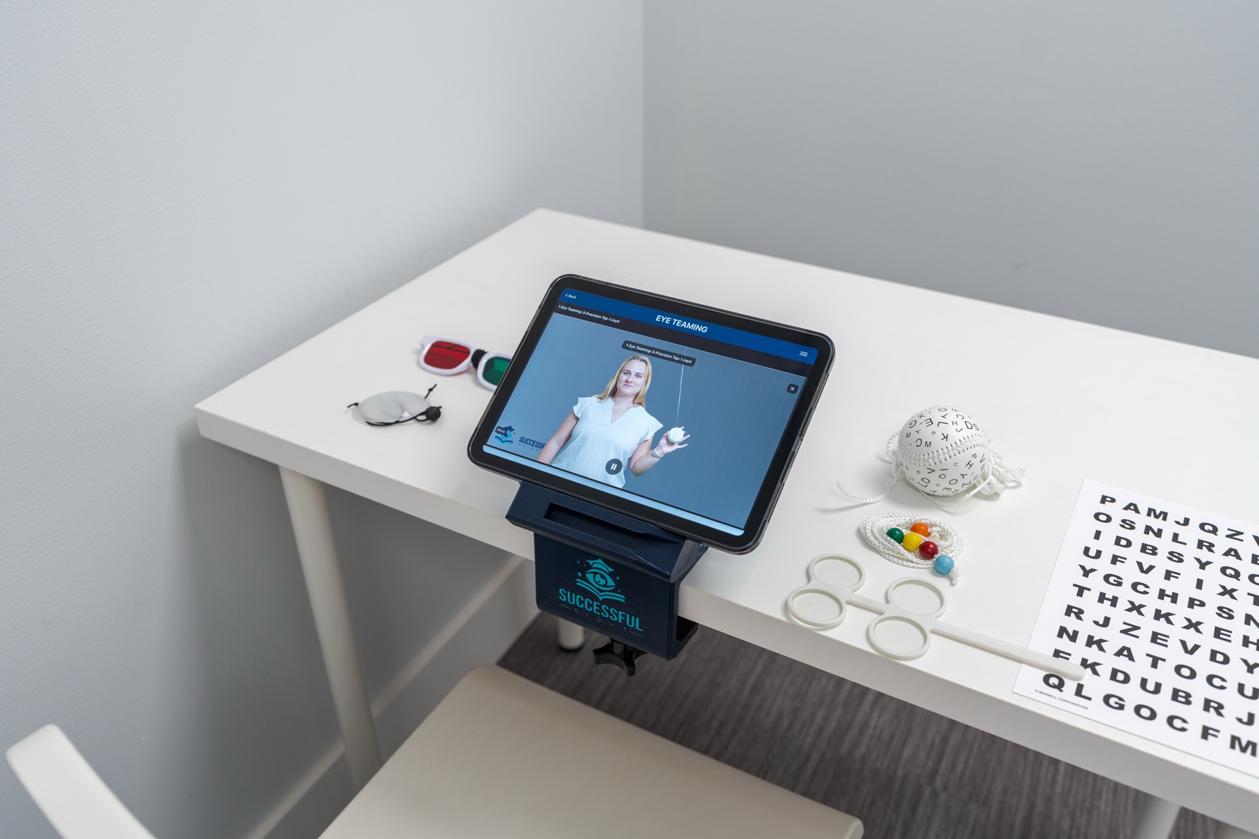 A patient's iPad on the SuccessfulSight™ stand playing an Eye Teaming therapy video, with red-green glasses, an eye patch, a Marsden ball, a Brock string, lens flippers, and a letter chart on the desk